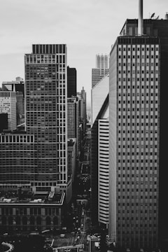 A black and white cityscape features towering skyscrapers lined along a narrow street. The buildings have varied architectural designs, including both modern glass exteriors and older brick facades. Street lights and a few vehicles are visible on the streets below.