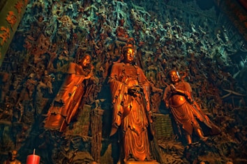 A detailed and intricate mural of religious or mythological figures, prominently featuring three large statues in the foreground with ornate robes and serene expressions. The background is densely filled with numerous smaller figures, adding a complex and layered texture to the scene.