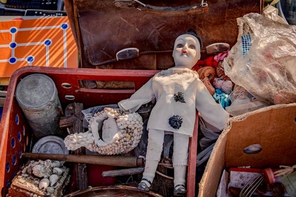 A collection of miscellaneous items is displayed, including an old doll with a painted face and white outfit, a ceramic ornament with seashells, a rustic canister, and a partially opened suitcase containing various objects like spools of thread and plastic bags.