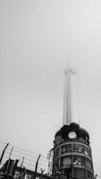 A tall communication tower reaches into a foggy sky. The structure features various antennas and circular installations on its cylindrical surface. The lower section is surrounded by a fence topped with barbed wire, suggesting restricted access.