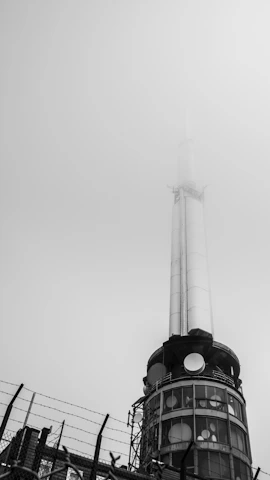 A tall communication tower reaches into a foggy sky. The structure features various antennas and circular installations on its cylindrical surface. The lower section is surrounded by a fence topped with barbed wire, suggesting restricted access.