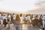 A large group of people gathered at a scenic overlook during sunset, capturing photos and enjoying the view. The atmosphere is lively with tourists engaged in various activities. The skyline and landscape stretch into the distance under a clear sky.