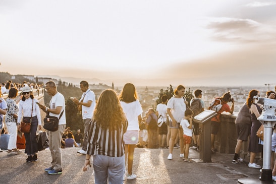 A large group of people gathered at a scenic overlook during sunset, capturing photos and enjoying the view. The atmosphere is lively with tourists engaged in various activities. The skyline and landscape stretch into the distance under a clear sky.