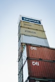 A tall stack of shipping containers with various labels such as 'FREITAG', 'SHOP', 'ZÜRICH', and 'CAI' against a clear sky backdrop. The containers are stacked in a way that creates a tower-like structure.