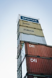 A tall stack of shipping containers with various labels such as 'FREITAG', 'SHOP', 'ZÜRICH', and 'CAI' against a clear sky backdrop. The containers are stacked in a way that creates a tower-like structure.