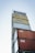 A tall stack of shipping containers with various labels such as 'FREITAG', 'SHOP', 'ZÜRICH', and 'CAI' against a clear sky backdrop. The containers are stacked in a way that creates a tower-like structure.