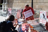 A street protest scene with an older man engaging in an animal rights demonstration. He is wearing a sign that reads 'I am an animal, don't kill me, I want to live' and is surrounded by various posters advocating for animal rights, veganism, and against animal cruelty. A younger person appears to be signing a petition or engaging in a conversation. The backdrop is a stone building with metal barricades lining the area.