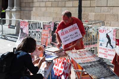 A street protest scene with an older man engaging in an animal rights demonstration. He is wearing a sign that reads 'I am an animal, don't kill me, I want to live' and is surrounded by various posters advocating for animal rights, veganism, and against animal cruelty. A younger person appears to be signing a petition or engaging in a conversation. The backdrop is a stone building with metal barricades lining the area.