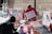 A street protest scene with an older man engaging in an animal rights demonstration. He is wearing a sign that reads 'I am an animal, don't kill me, I want to live' and is surrounded by various posters advocating for animal rights, veganism, and against animal cruelty. A younger person appears to be signing a petition or engaging in a conversation. The backdrop is a stone building with metal barricades lining the area.