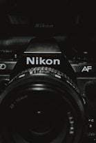 A close-up view of a Nikon camera, focusing on the camera lens and the brand name prominently displayed. The image is dark with lighting that highlights the details of the camera body and lens, creating a dramatic effect.