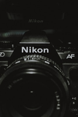 A close-up view of a Nikon camera, focusing on the camera lens and the brand name prominently displayed. The image is dark with lighting that highlights the details of the camera body and lens, creating a dramatic effect.