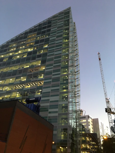 A sleek, modern skyscraper under construction at dusk, illuminated by warm golden lights against a dark sky.