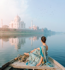 A person in a colorful sari is sitting on a boat, gently gliding on a calm river. The majestic Taj Mahal stands in the distance, reflected beautifully in the water. A flock of birds is flying overhead, adding to the serene atmosphere, and the sky appears partly cloudy with soft hues.