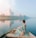 A person in a colorful sari is sitting on a boat, gently gliding on a calm river. The majestic Taj Mahal stands in the distance, reflected beautifully in the water. A flock of birds is flying overhead, adding to the serene atmosphere, and the sky appears partly cloudy with soft hues.