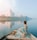 A person in a colorful sari is sitting on a boat, gently gliding on a calm river. The majestic Taj Mahal stands in the distance, reflected beautifully in the water. A flock of birds is flying overhead, adding to the serene atmosphere, and the sky appears partly cloudy with soft hues.