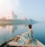 A person in a colorful sari is sitting on a boat, gently gliding on a calm river. The majestic Taj Mahal stands in the distance, reflected beautifully in the water. A flock of birds is flying overhead, adding to the serene atmosphere, and the sky appears partly cloudy with soft hues.