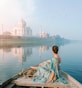 A person in a colorful sari is sitting on a boat, gently gliding on a calm river. The majestic Taj Mahal stands in the distance, reflected beautifully in the water. A flock of birds is flying overhead, adding to the serene atmosphere, and the sky appears partly cloudy with soft hues.