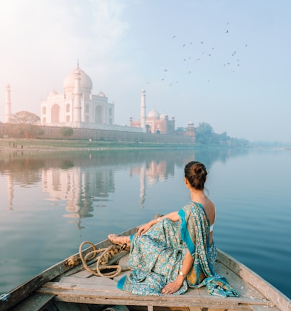 A person in a colorful sari is sitting on a boat, gently gliding on a calm river. The majestic Taj Mahal stands in the distance, reflected beautifully in the water. A flock of birds is flying overhead, adding to the serene atmosphere, and the sky appears partly cloudy with soft hues.
