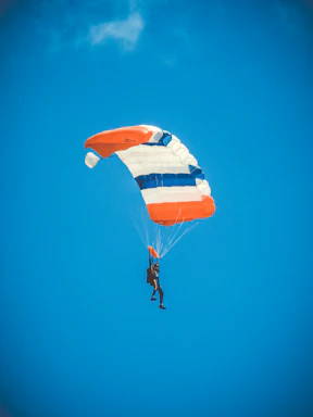 A skydiver in freefall with a vibrant orange parachute against a clear blue sky.