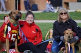 Two women sitting on grass with two dogs. The woman on the left is wearing a red jacket, and the large dog beside her is wearing a Maryland sports jersey. The woman on the right is wearing sunglasses and a dark jacket, holding a smaller dog on a leash.