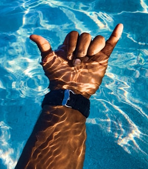 A hand is making a 'shaka' gesture, partially submerged in clear blue water with sunlight creating intricate patterns on the skin. The hand wears a dark wristband.