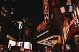 A nighttime urban scene filled with vibrant neon signs and illuminated advertisements. Prominent signage includes Regal Cinemas and various restaurants and shops. The atmosphere is bustling with bright lights against a dark city backdrop.