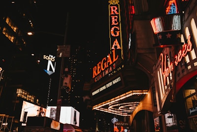 A nighttime urban scene filled with vibrant neon signs and illuminated advertisements. Prominent signage includes Regal Cinemas and various restaurants and shops. The atmosphere is bustling with bright lights against a dark city backdrop.