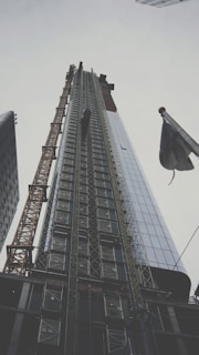 A towering skyscraper is under construction, its structure framed by scaffolding and a tall crane on the left. The building's glass facade reflects the cloudy sky, while an adjacent flag flutters in the foreground.