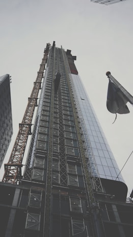 A towering skyscraper is under construction, its structure framed by scaffolding and a tall crane on the left. The building's glass facade reflects the cloudy sky, while an adjacent flag flutters in the foreground.