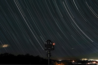 A long exposure photograph capturing star trails in the night sky. The trails form graceful arcs across the image, suggesting the Earth's rotation. Below the celestial display, a silhouette of a camera on a tripod is visible, indicating the scene was carefully composed. The lower part of the image shows a faint horizon with dark silhouettes of trees and distant lights.