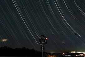 A long exposure photograph capturing star trails in the night sky. The trails form graceful arcs across the image, suggesting the Earth's rotation. Below the celestial display, a silhouette of a camera on a tripod is visible, indicating the scene was carefully composed. The lower part of the image shows a faint horizon with dark silhouettes of trees and distant lights.