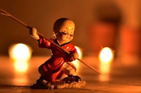 A small figurine of a martial artist in a dynamic pose, wearing a red robe and holding a staff. The background features warm glowing lights, giving a sense of depth and ambiance.