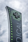 A tall, modern sign with a dark background featuring a large, illuminated green cross at the top. The word 'MEDICAL' is written vertically in large letters. Smaller green crosses line the side of the sign, set against a cloudy sky.