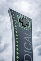 A tall, modern sign with a dark background featuring a large, illuminated green cross at the top. The word 'MEDICAL' is written vertically in large letters. Smaller green crosses line the side of the sign, set against a cloudy sky.