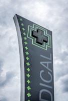 A tall, modern sign with a dark background featuring a large, illuminated green cross at the top. The word 'MEDICAL' is written vertically in large letters. Smaller green crosses line the side of the sign, set against a cloudy sky.