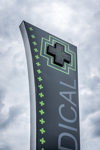 A tall, modern sign with a dark background featuring a large, illuminated green cross at the top. The word 'MEDICAL' is written vertically in large letters. Smaller green crosses line the side of the sign, set against a cloudy sky.