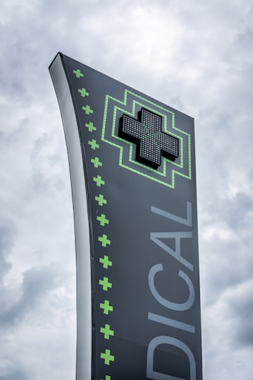 A tall, modern sign with a dark background featuring a large, illuminated green cross at the top. The word 'MEDICAL' is written vertically in large letters. Smaller green crosses line the side of the sign, set against a cloudy sky.