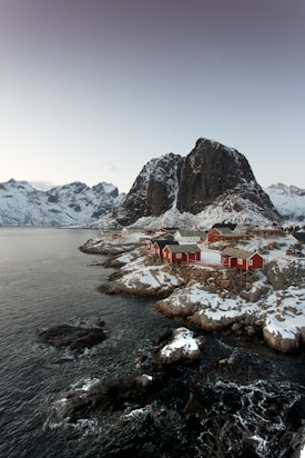A coastal landscape featuring several red wooden cabins situated on rocky terrain by the sea. Snow covers parts of the ground and rooftops, while large, rugged mountains rise dramatically in the background. The calm water and soft light create a serene and tranquil atmosphere.