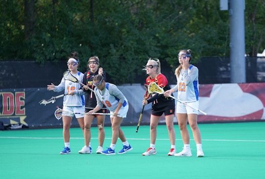 Five people are standing on a sports field with green artificial turf. Each person is wearing protective eyewear and holding a lacrosse stick. They are dressed in athletic gear, with a mix of white, blue, orange, and black colors. The background features a fence and some trees.