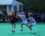 A group of women are playing lacrosse on a green field. Two players in light-colored uniforms are attempting to take possession of the ball from a player in a black and red uniform. The background shows a tent and a line of people observing the game.