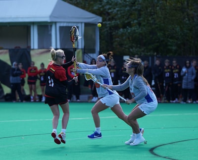 A group of women are playing lacrosse on a green field. Two players in light-colored uniforms are attempting to take possession of the ball from a player in a black and red uniform. The background shows a tent and a line of people observing the game.