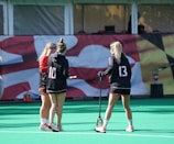Three people are seen on a sports field, likely preparing for a field hockey or lacrosse game, given the presence of a stick. They are wearing sports attire, including jerseys with numbers, and are engaged in conversation. A banner with a graphic design serves as the background.