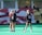 Three people are seen on a sports field, likely preparing for a field hockey or lacrosse game, given the presence of a stick. They are wearing sports attire, including jerseys with numbers, and are engaged in conversation. A banner with a graphic design serves as the background.