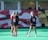 Three people are seen on a sports field, likely preparing for a field hockey or lacrosse game, given the presence of a stick. They are wearing sports attire, including jerseys with numbers, and are engaged in conversation. A banner with a graphic design serves as the background.