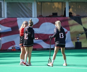 Three people are seen on a sports field, likely preparing for a field hockey or lacrosse game, given the presence of a stick. They are wearing sports attire, including jerseys with numbers, and are engaged in conversation. A banner with a graphic design serves as the background.