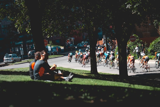 A group of cyclists is participating in a race on a road surrounded by trees and spectators. Several people are sitting on a grassy hill in the foreground, watching the race. The cyclists are wearing colorful jerseys and helmets. The scene takes place in an urban setting with parked cars in the background.
