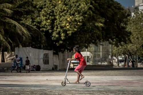 A young child rides a scooter across an open paved area, with trees providing shade in the background. The child wears a bright red shirt and pink shorts, and there are other people in the distance, one of whom appears to be riding a bicycle. The setting has a casual and relaxed atmosphere.