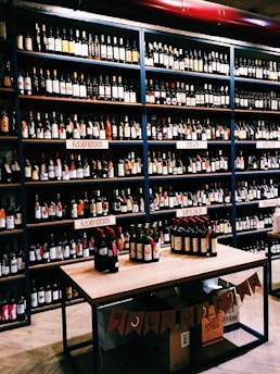 Shelves filled with various bottles of wine displayed in a store. The labels on the bottles and the signs above the shelves suggest a diverse selection. A table in the foreground exhibits additional bottles, and boxes are stacked below it with banners hanging down.