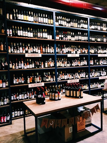 Shelves filled with various bottles of wine displayed in a store. The labels on the bottles and the signs above the shelves suggest a diverse selection. A table in the foreground exhibits additional bottles, and boxes are stacked below it with banners hanging down.