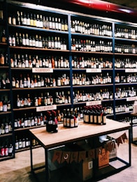 Shelves filled with various bottles of wine displayed in a store. The labels on the bottles and the signs above the shelves suggest a diverse selection. A table in the foreground exhibits additional bottles, and boxes are stacked below it with banners hanging down.